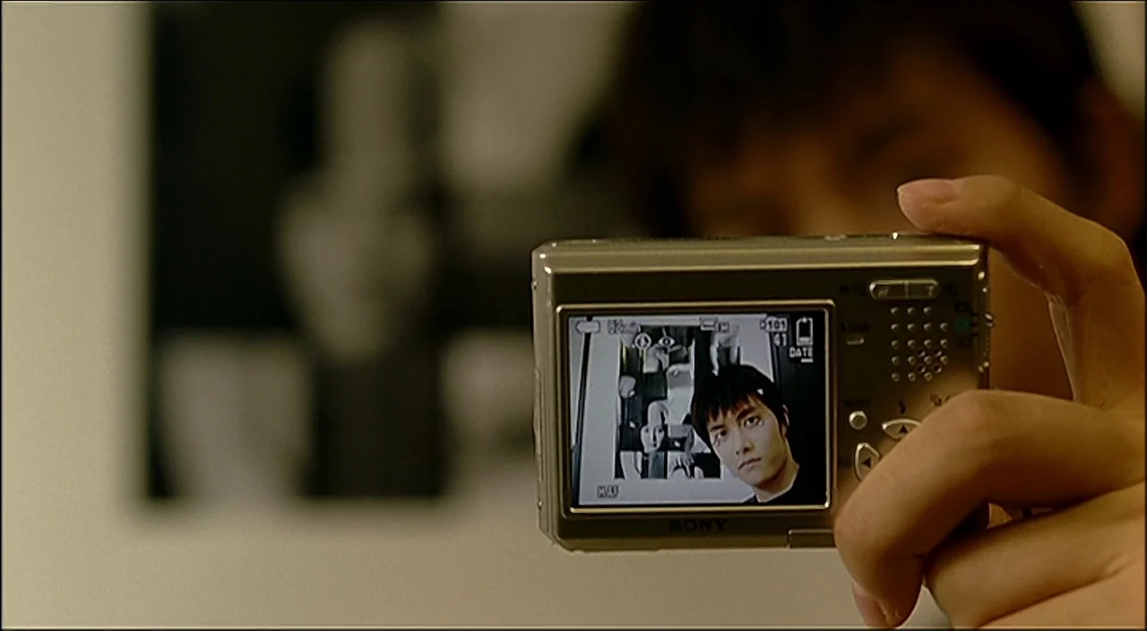 3-Iron (2004) movie still: closeup — A close view shows a hand holding a silver digital camera towards the viewer. The camera screen displays a…