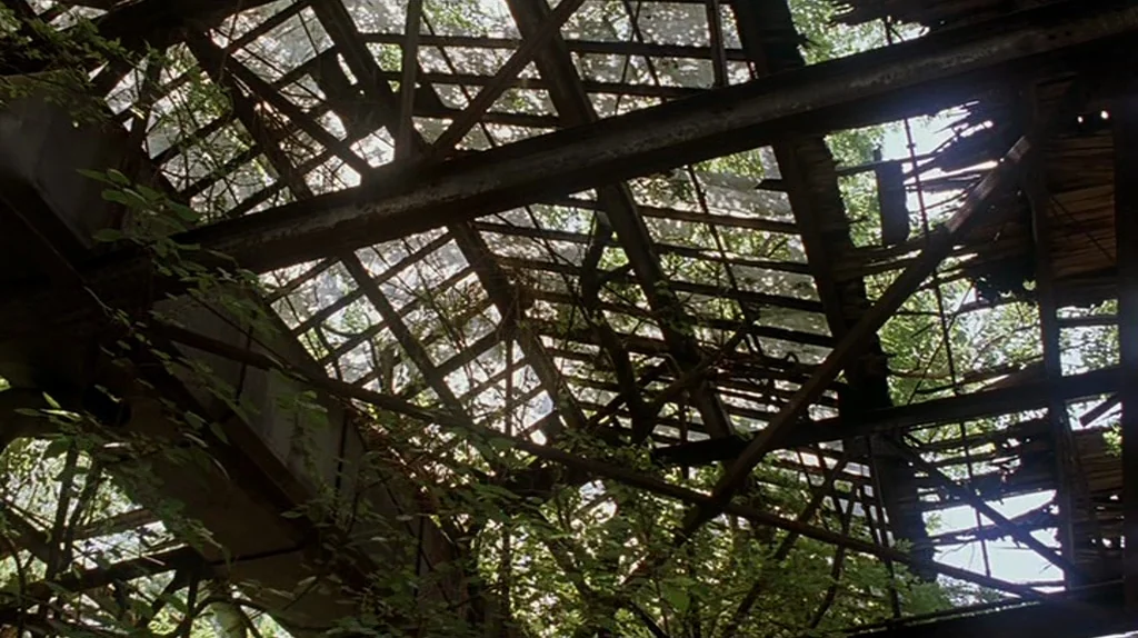 Undertow (2004) movie still: establishing — A dilapidated greenhouse features a collapsed glass ceiling supported by rusted iron girders. Lush green…