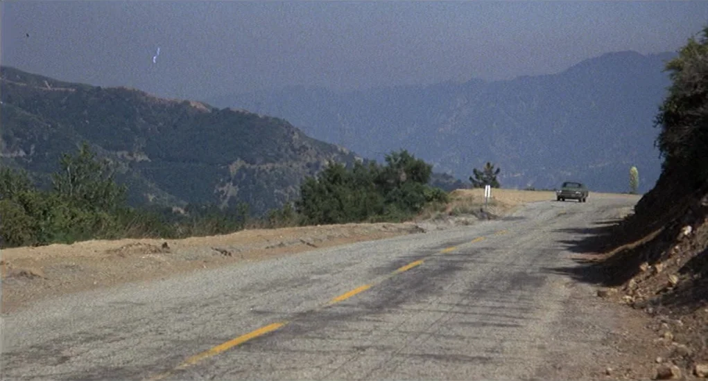 Family Plot (1976) movie still: wide — An asphalt road winds through a dry, rugged mountain landscape under a clear sky. A single sedan travels…