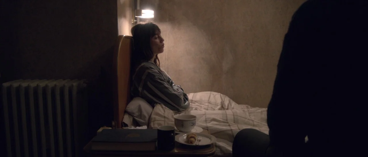Nymphomaniac Vol 1 (2013) movie still: medium — A dim room features a woman sitting in bed near a radiator and nightstand. The foreground is…