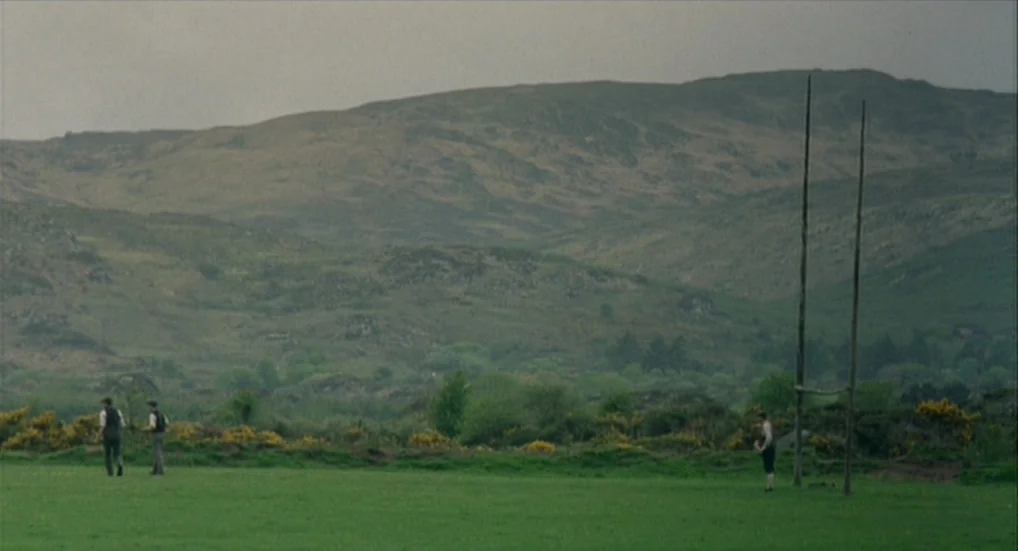 The Wind That Shakes the Barley (2006) movie still: wide — A rural Irish landscape features rolling hills behind a vast, open green field. Three men…