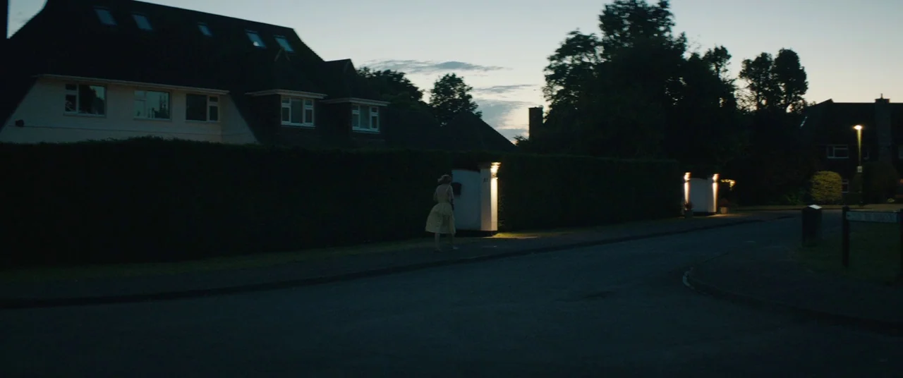 Beast (2017) movie still: wide — A quiet residential street at dusk is flanked by tall, dark trimmed hedges. A lone woman in a yellow dress walks…