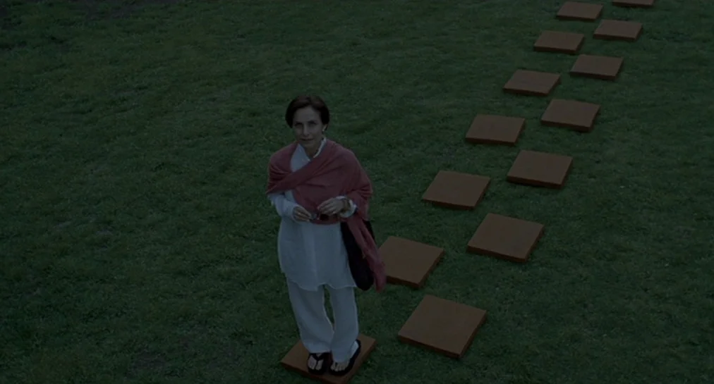 Y Tu Mamá También (2001) movie still: fullbody — A dark green grassy lawn features a path of square stone pavers. A woman stands on one of the…