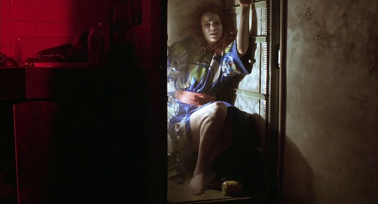 Hardware (1990) movie still: medium — A woman crouches inside a cramped, dark closet illuminated by a harsh light. She holds the edge of the door…