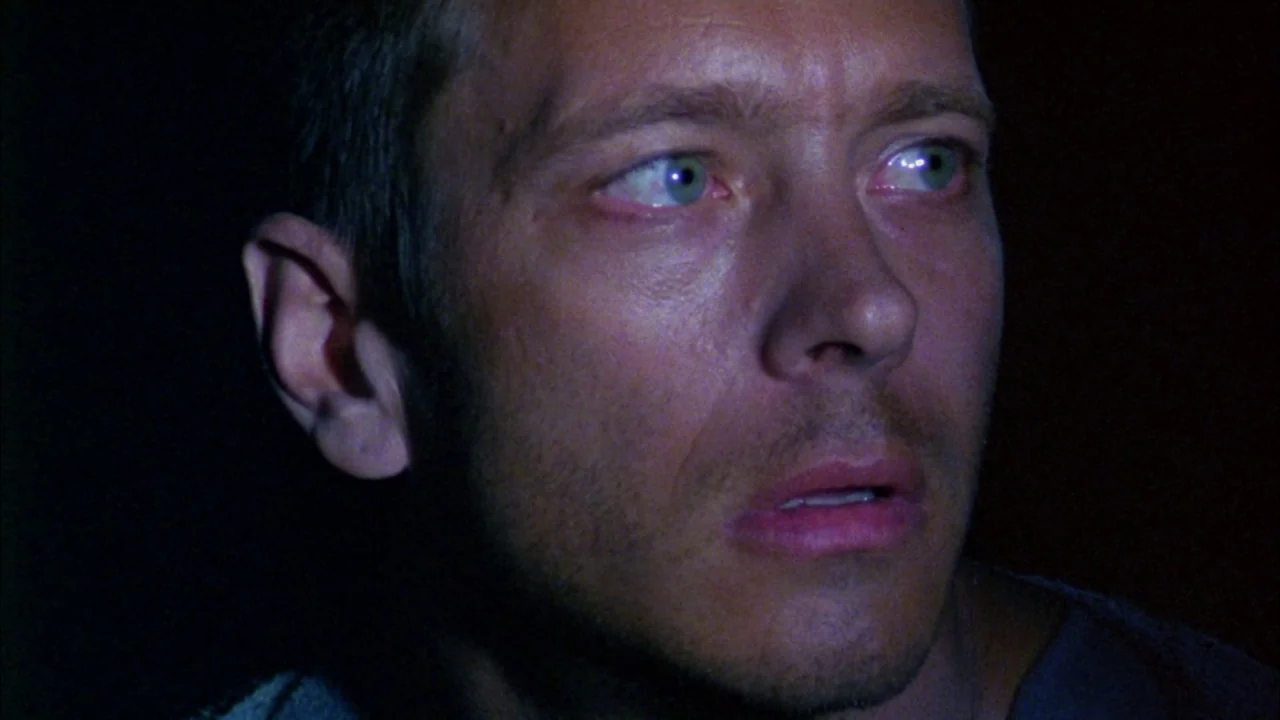 Poison movie still: closeup — A close portrait of a man's face set against a pitch black background. The man gazes forward with wide eyes as if…