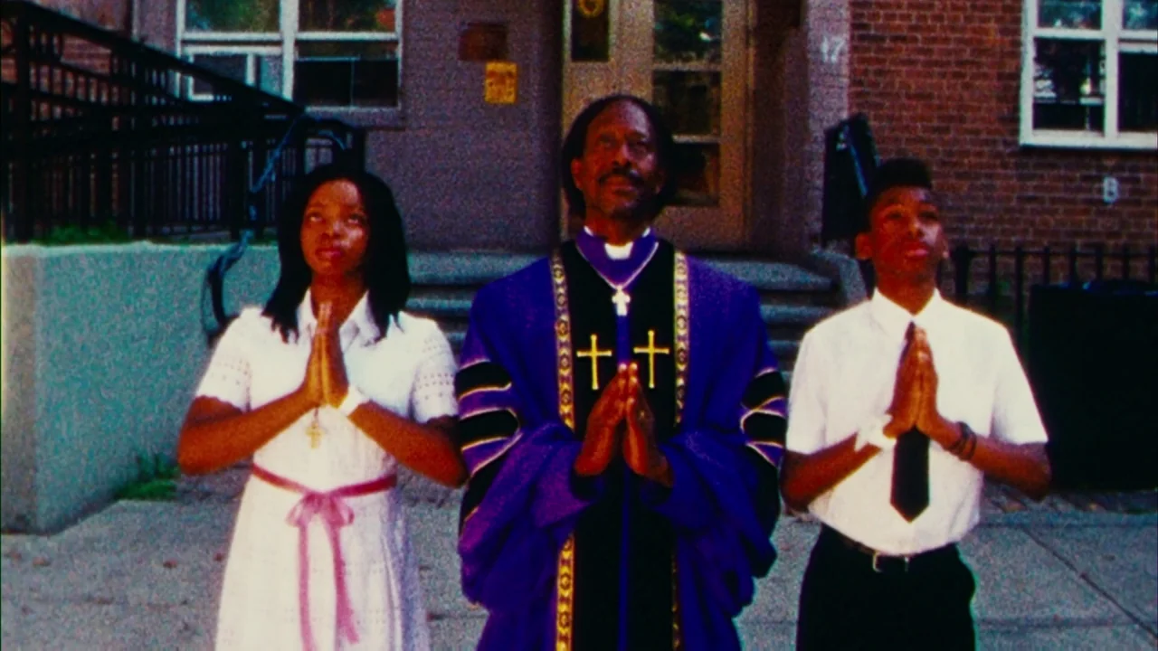 Red Hook Summer (2012) movie still: fullbody — Three people stand in prayer before a brick institutional building with glass doors. A girl, an older…