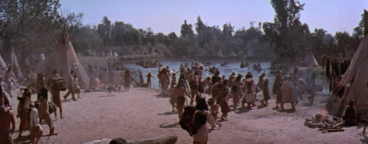 The Seven Year Itch (1955) movie still: wide — An indigenous encampment sits along a riverbank with multiple conical teepees and people gathered.…