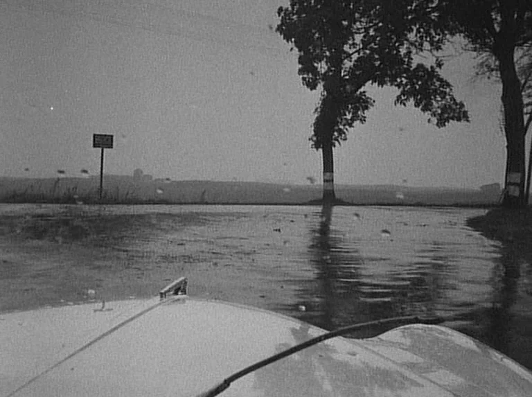 Knife In The Water (1962) movie still: wide — A wet asphalt road stretches toward a distant tree line under an overcast sky. A car hood with a…