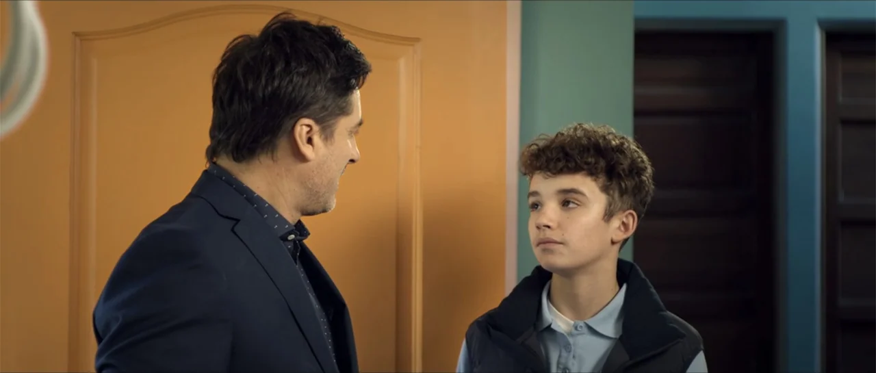 Disconnected 2 (2025) movie still: medium — An interior hallway features a tan door and a teal wall with recessed dark panels. An adult man in a…