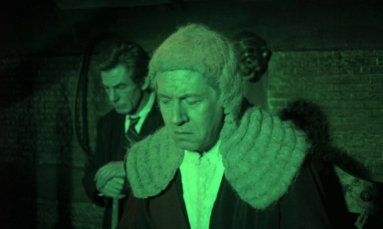 Curse of the Crimson Altar (1968) movie still: medium — A dark, brick-walled interior resembles a dimly lit courtroom during a night trial. The…
