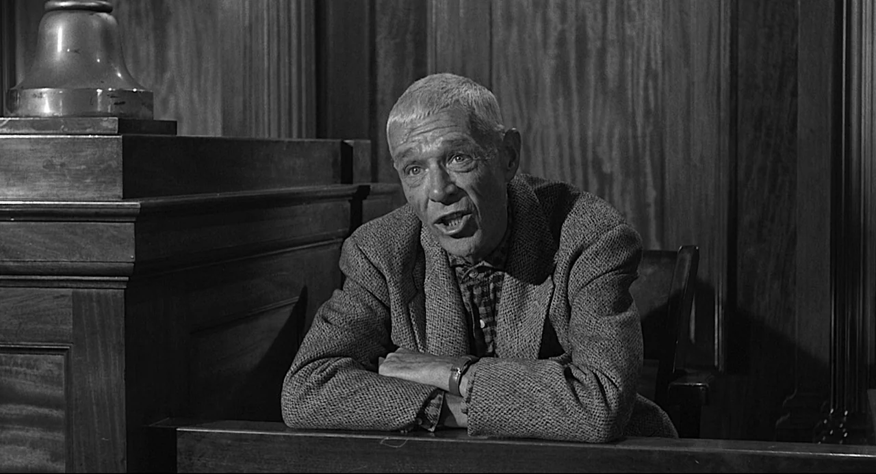 Anatomy of a Murder (1959) movie still: medium — An older man sits on a wooden witness stand in a formal room. He rests his crossed arms on the…