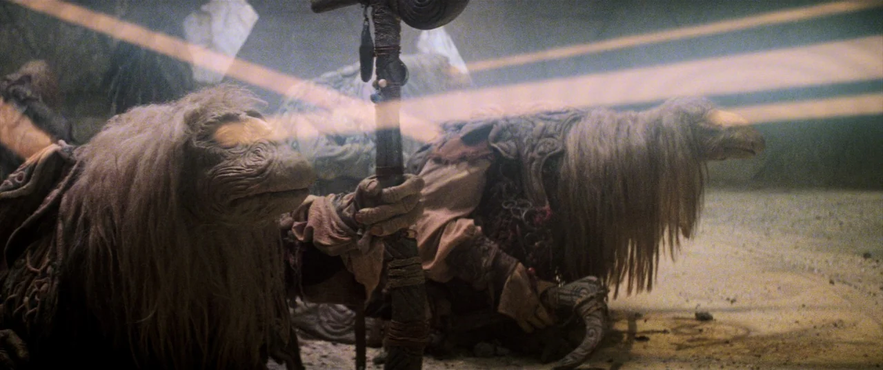 The Dark Crystal (1982) movie still: medium — A dark, rocky interior space illuminated by a thin, glowing horizontal beam of light. Two shaggy,…
