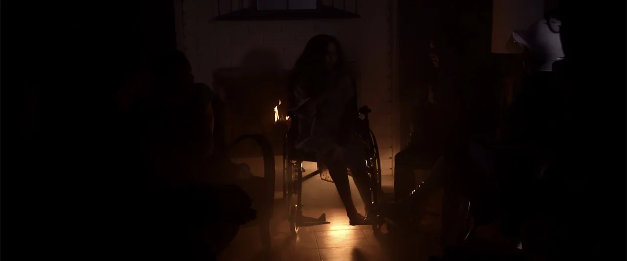 Mermaid Down (2019) movie still: medium — A dark living room centered around a fireplace with several silhouettes present. A woman sits in a…