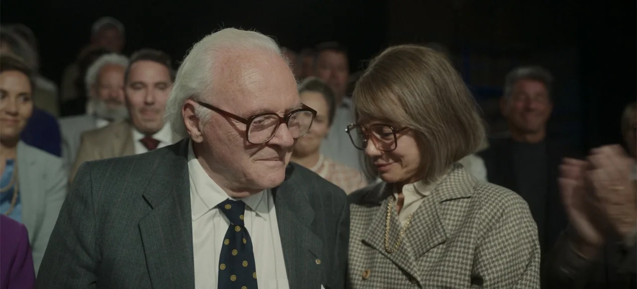 One Life (2023) movie still: medium — A crowded indoor event space filled with people clapping at an awards ceremony. An elderly man and woman stand…