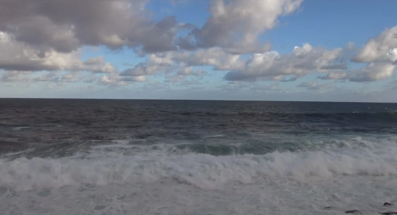A Dramatic Film (2019) movie still: establishing — A vast ocean view under a blue sky filled with scattered white clouds. Rough waves break in the…