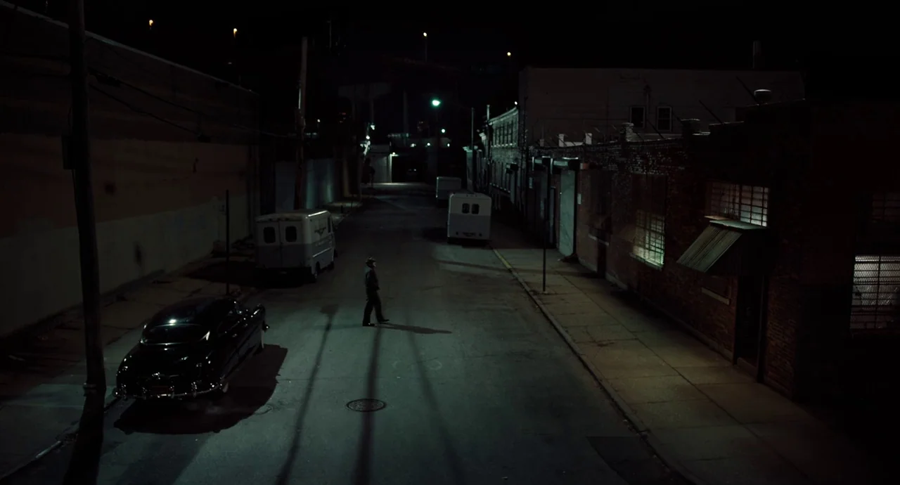 The Irishman (2019) movie still: wide — A quiet urban side street at night features brick walls and parked service vans. A lone man walks across the…