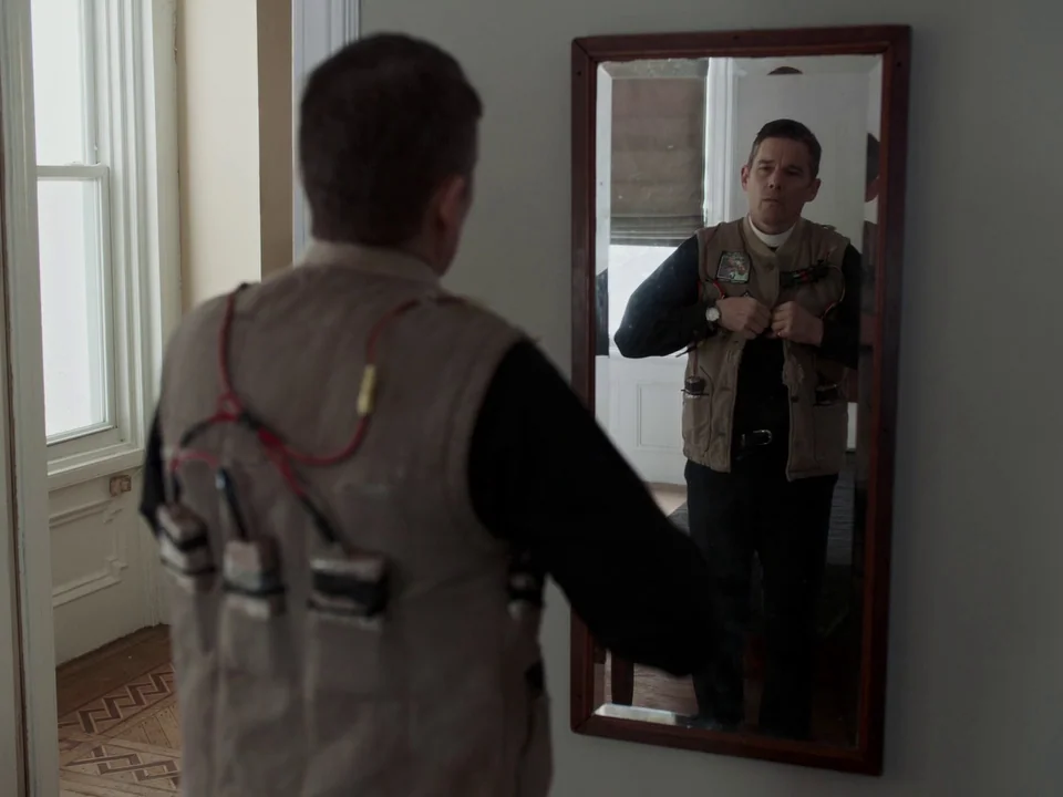 First Reformed (2017) movie still: medium — A man stands in a hallway facing a mirror hanging on a gray wall. The man wearing a beige vest with…