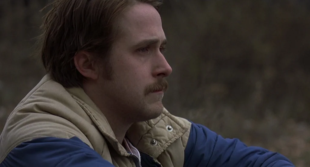 Lars And The Real Girl (2007) movie still: closeup — A man stands in an outdoor area with blurred, natural textures in the distance. The person with…