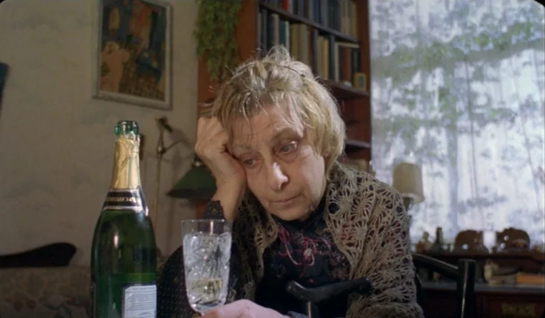 The Turn of the Century (2001) movie still: medium — A cluttered room contains a bookshelf and a table with a glass of wine. An elderly woman sits…