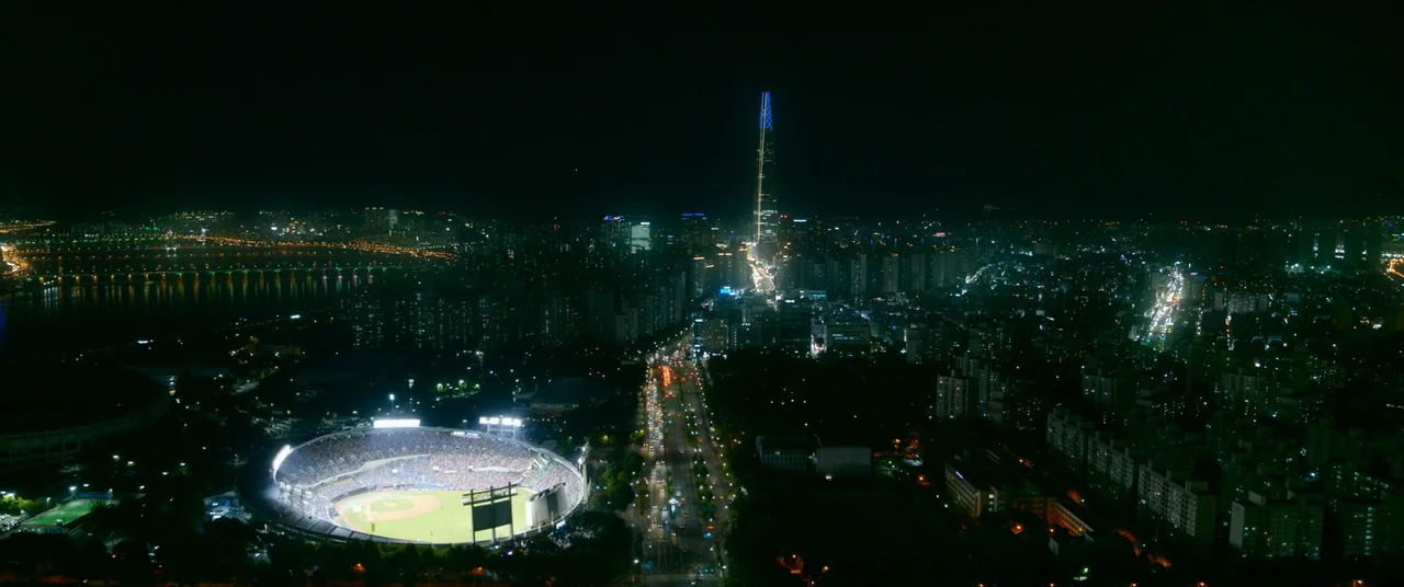 The Divine Fury (2019) movie still: establishing — An aerial night view of a dense metropolitan area under a dark sky. A bright stadium with a…