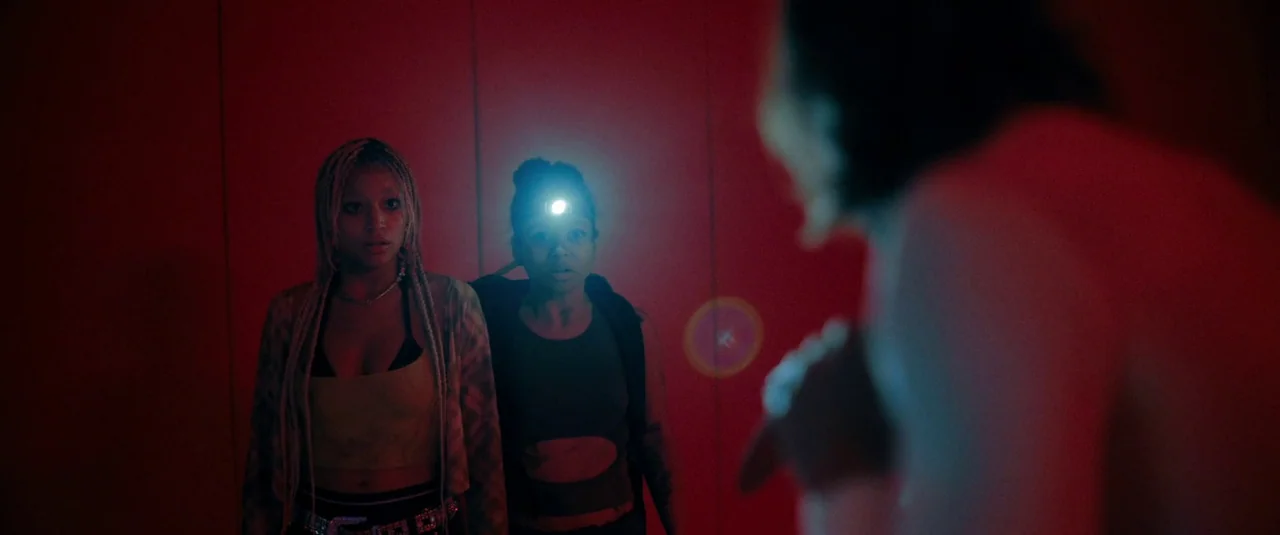 Bodies Bodies Bodies (2022) movie still: medium — Two women stand in a dark, red-lit corridor with smooth vertical wall panels. They look toward an…