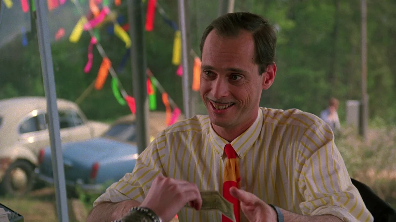 Something Wild (1986) movie still: medium — An outdoor area is decorated with colorful ribbons hanging from a light metal structure. A man with a…