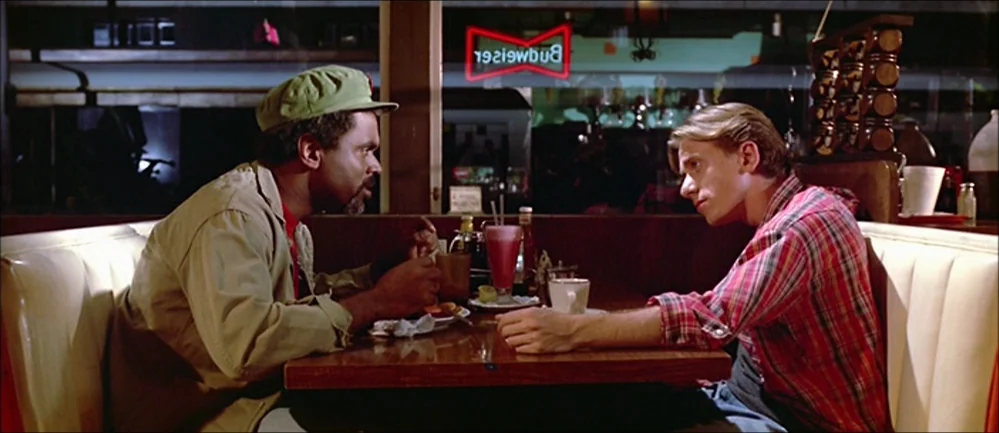 Reservoir Dogs (1992) movie still: medium — Two men sit across from each other in a vinyl-upholstered booth at night. A red neon sign is visible…