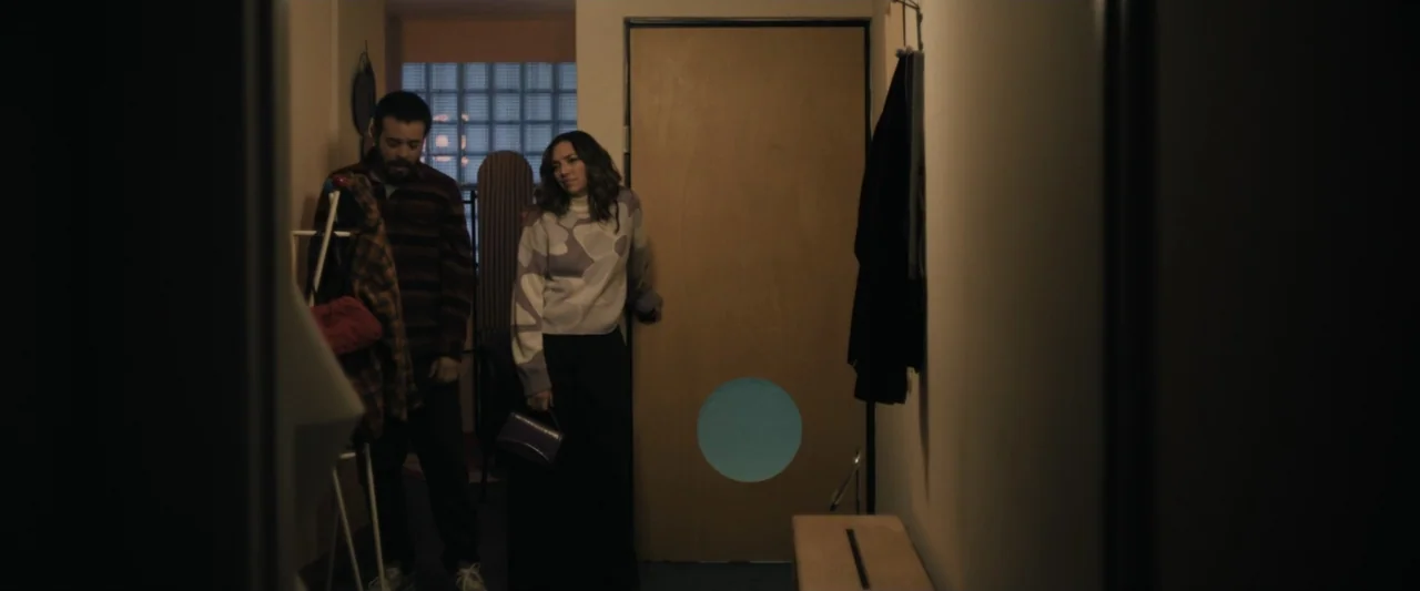 Sign Here (2017) movie still: medium — A dimly lit narrow apartment hallway with a wooden door featuring a blue circle. A man and a woman stand…