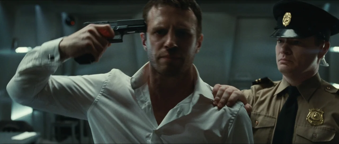 Slackers (2002) movie still: medium — A dimly lit room with metallic surfaces and sterile industrial aesthetics. A man in a white shirt points a…