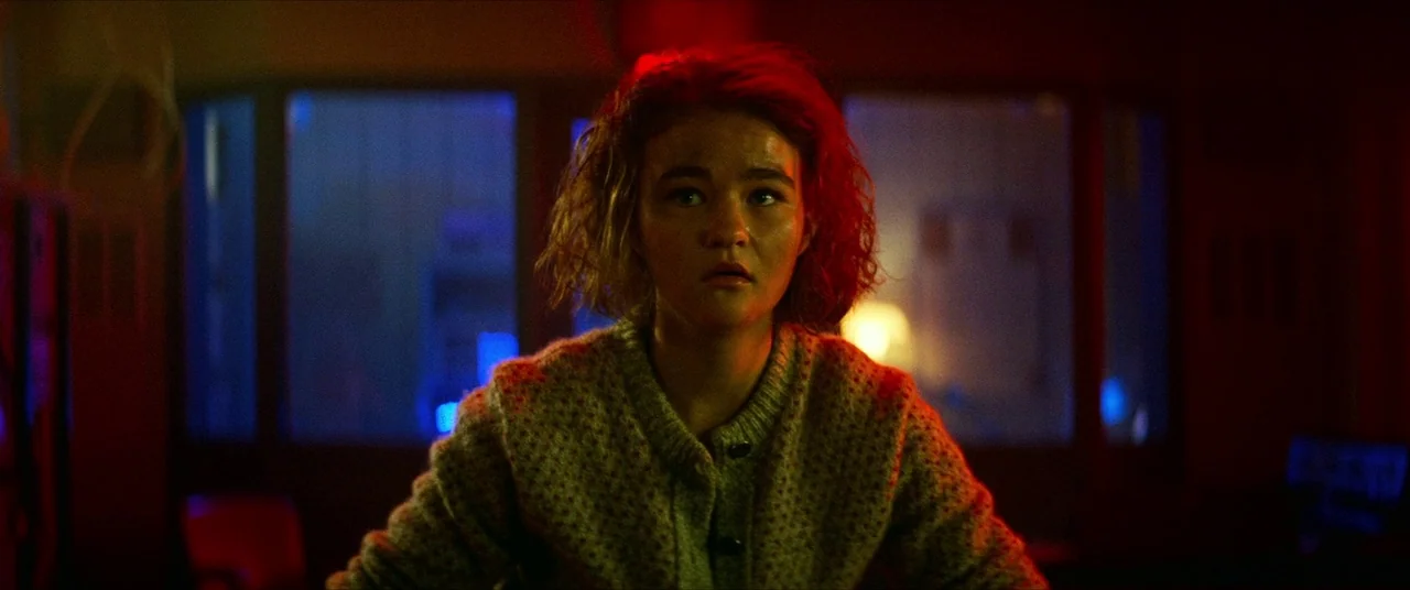 A Quiet Place Part II (2020) movie still: medium — A dimly lit office interior with glass panels reflected in the background. A young woman stands…