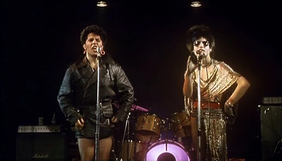 Labyrinth of Passion (1982) movie still: medium — A dimly lit musical performance space with a black backdrop and drum kit. Two singers with…
