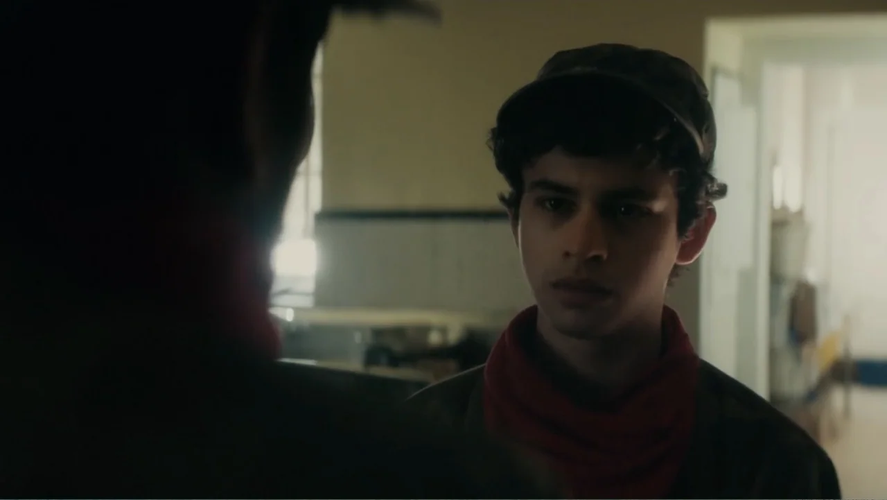 Bel Canto (2018) movie still: medium — An interior space features pale walls and a blurred doorway in the background. A young man wearing a red…