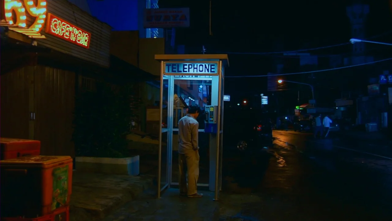 Merantau (2009) movie still: wide — A dark city street at night features a lone glass telephone booth on the sidewalk. A man stands inside the booth…
