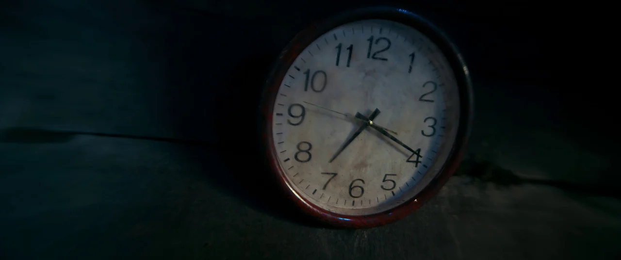 One And Four (2021) movie still: closeup — A close-up of a circular analog clock resting on a dark surface in shadow. The clock face shows the time…