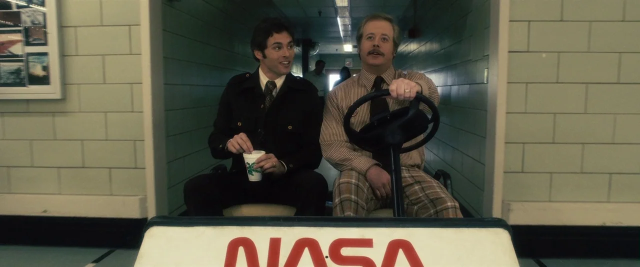 The Box (2009) movie still: medium — A long institutional hallway with light green brick walls is shown. Two men sit in a white NASA golf cart…