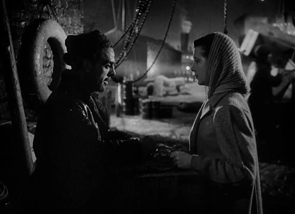 Odd Man Out (1947) movie still: medium — A dark, industrial maritime setting with heavy ropes and nautical rigging in the background. A man in a…
