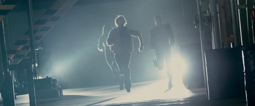 MacGruber (2010) movie still: fullbody — An industrial facility with metal piping and dark barrels along the side walls. Three people run toward the…