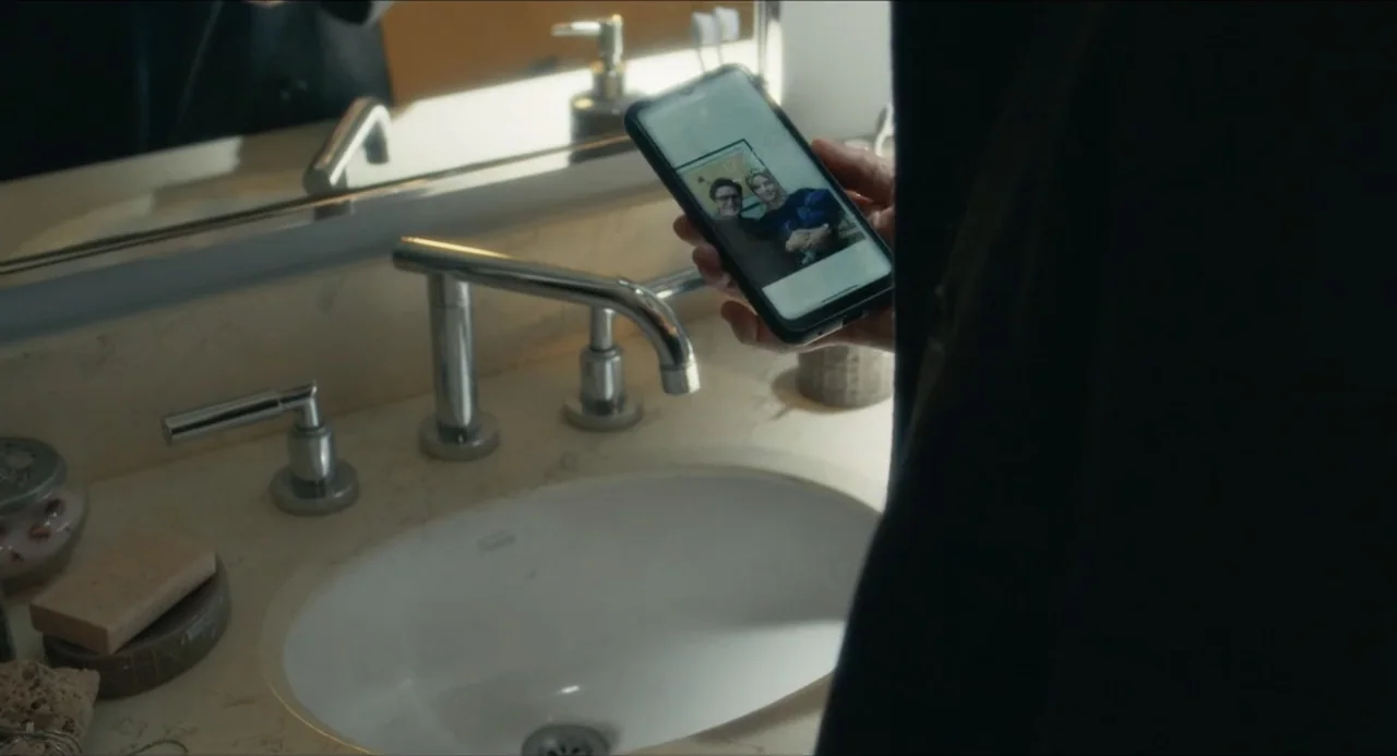 Echoes Of A Crime (2025) movie still: medium — A modern bathroom features a beige vanity top with a chrome faucet and sink. A person stands facing…