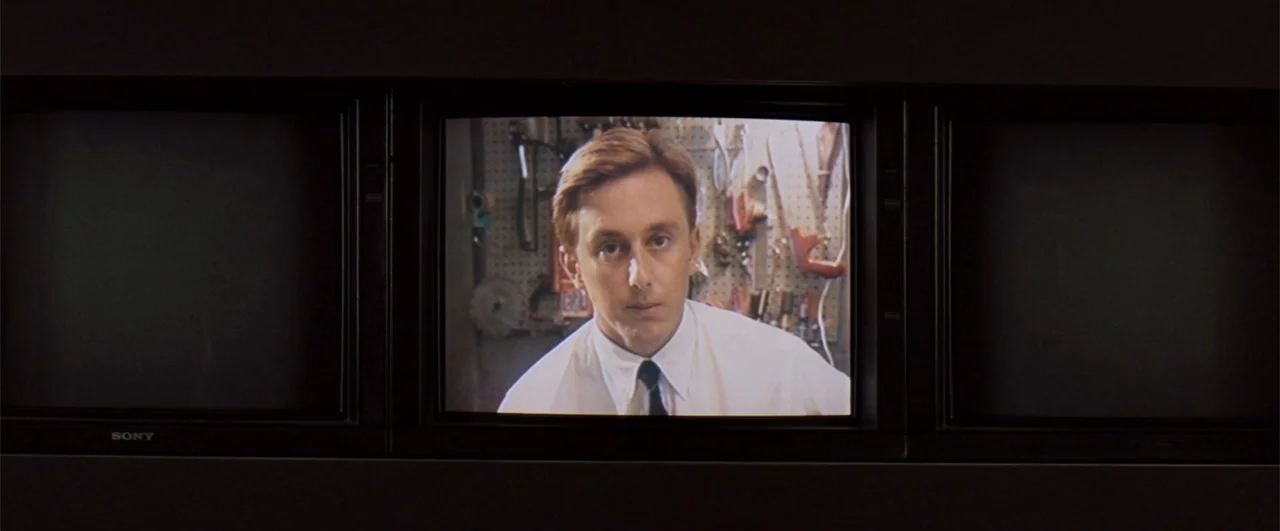 The Pelican Brief (1993) movie still: medium — A man appears on a television monitor screen in a dark room. The screen shows the man in front of a…