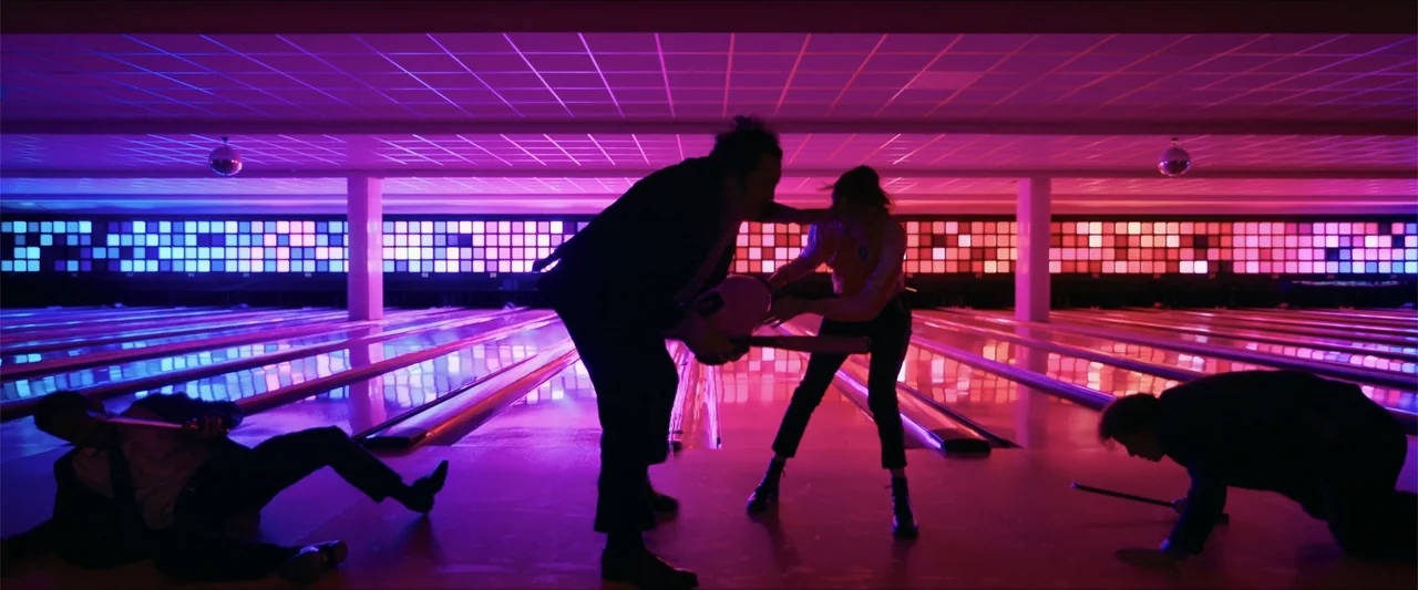 Gunpowder Milkshake (2021) movie still: fullbody — A dark, saturated bowling alley is illuminated by vibrant magenta and blue grid panels. Four…
