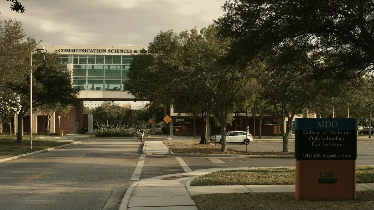 Death of a Child (2017) movie still: establishing — A university campus entrance features a large brick bridge building and signage for MDO. The…