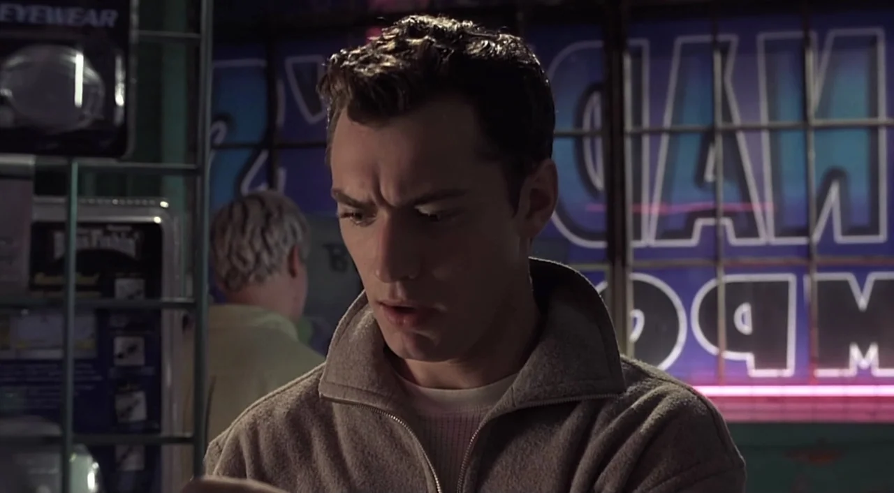eXistenZ (1999) movie still: medium — A retail setting features shelves with packaged products and a large blue neon sign. A man in a fleece jacket…