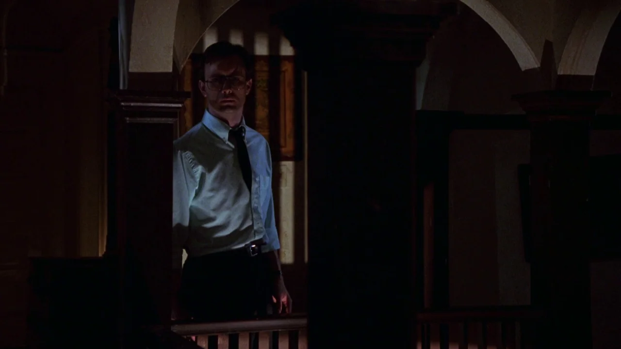 Bride of Reanimator (1989) movie still: medium — A dimly lit residential hallway features ornate wooden banisters and arched architectural details.…