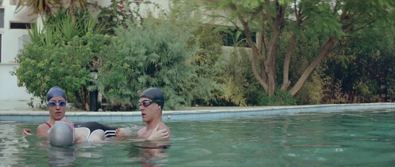Dogtooth movie still: medium — Two swimmers support an unresponsive person in the water next to a stone poolside. The area is surrounded by lush…