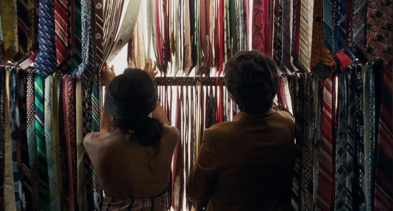 Inherent Vice (2014) movie still: medium — A dense wall of hanging neckties occupies a retail clothing space. A man and woman stand before the…