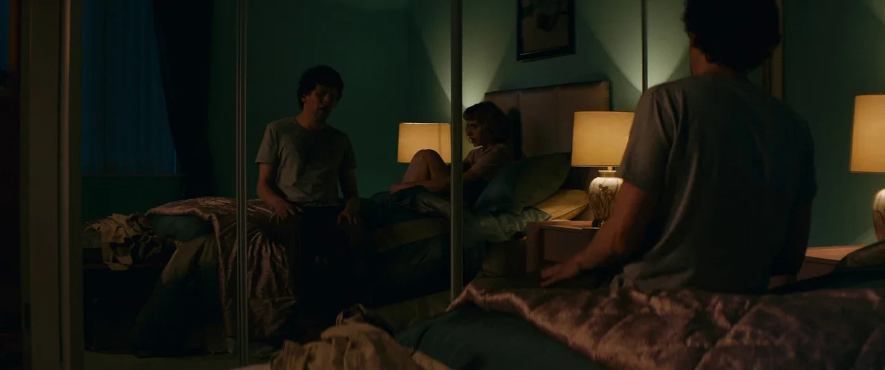 Vivarium (2019) movie still: wide — A dimly lit room features a bed positioned in front of a mirrored closet door. A man sits on the edge of the…