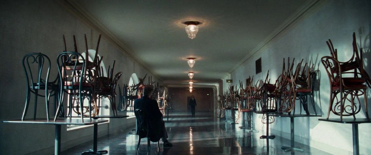 Steve Jobs (2015) movie still: wide — A long, symmetrical hallway features high ceilings, light-colored walls, and a polished tiled floor. Chairs…