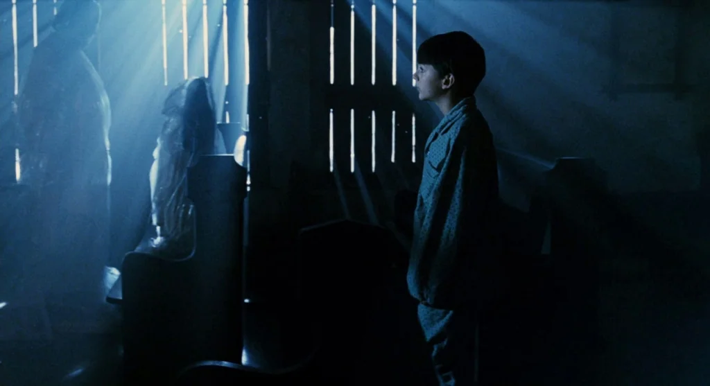 Mimic (1997) movie still: medium — A dark wooden structure features slats that allow beams of blue light inside. A young boy in pajamas stands in…