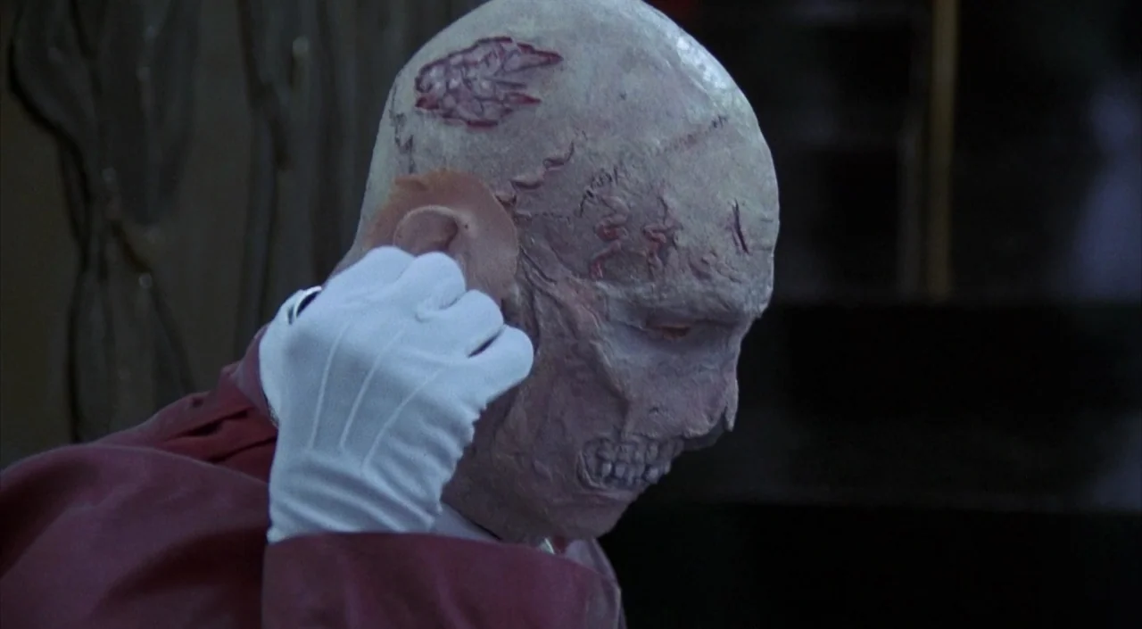 Dr. Phibes Rises Again (1972) movie still: closeup — A man applies a prosthetic ear to the side of his heavily scarred head. He wears a formal red…