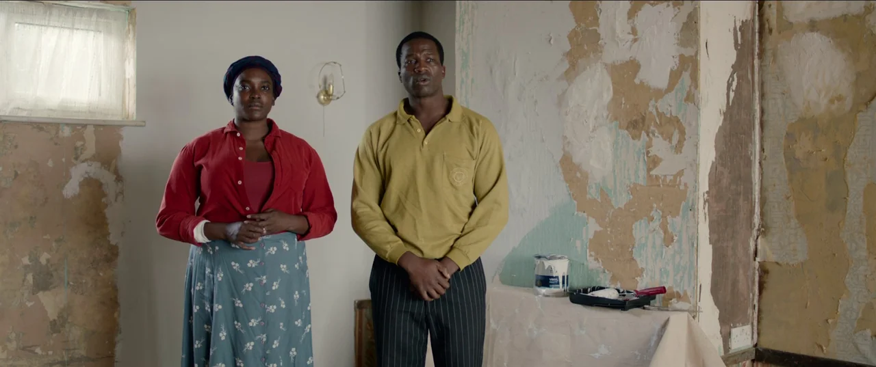 His House (2020) movie still: fullbody — A sparse room with peeling wallpaper and plaster patches on the walls during renovation. A man and a woman…